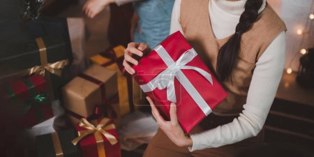 Foto de Mujer asiática joven con una banda de cabeza elegante sonríe y sostiene un regalo de Navidad en una caja roja con cinta en una sala de estar con un árbol de Navidad decorado en el fondo. - Imagen libre de derechos