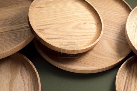 Flache Holzteller auf dunkelgrünem Hintergrund. Das Konzept des ökologischen Geschirrs. Produkte für die moderne Küche. Natürliche Harmonie: Holzplatten in einer umweltfreundlichen Küche. Warme Lampenschirme.