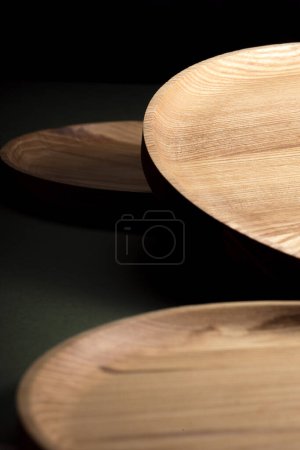 Flache Holzteller auf dunkelgrünem Hintergrund. Das Konzept des ökologischen Geschirrs. Produkte für die moderne Küche. Natürliche Harmonie: Holzplatten in einer umweltfreundlichen Küche. Warme Lampenschirme.