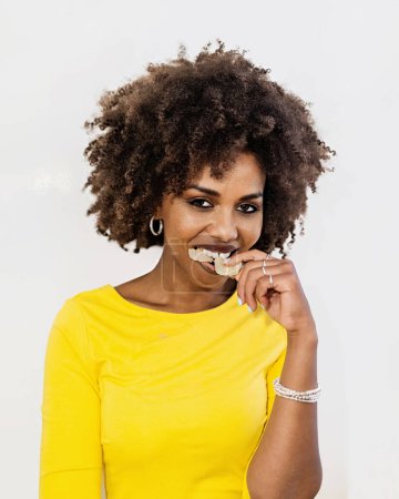 Photo for Headshot of curly haired African American 30s woman bites baked food winks looking at camera - a lady with yellow long sleeved shirt shirt eats unhealthy food poses indoor. People lifestyle concept - Royalty Free Image