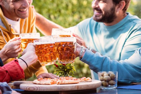 Photo for Cheerful group of young people clinking mood glasses over a sliced pizza - Millennial friends celebrating joining beer mugs at picnic outdoors in a sunny day - People gathering, food and drink lifestyle concept - Royalty Free Image