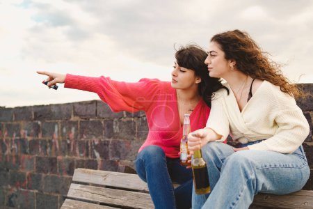 Téléchargez les photos : Two young girlfriends pointing away and drinking beers from bottles and sitting on a bench outdoors - people, food and drinks lifestyle concept - en image libre de droit