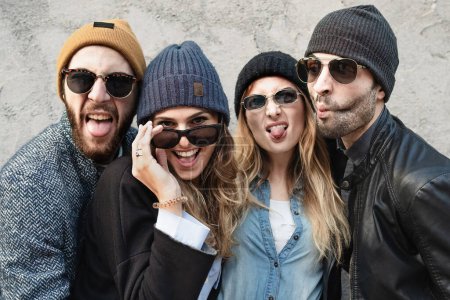 Foto de Portrait with millennials wearing sunglasses and wooden caps looking at the camera - Best friends posing isolated against a concrete wall making funny and weird faces - people lifestyle concept - Imagen libre de derechos