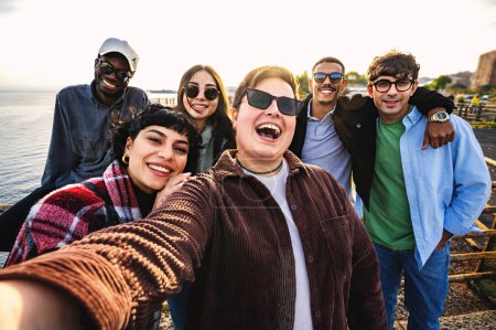 Téléchargez les photos : Group of happy multiethnic friends moment seaside - happy diverse group of friends enjoying a beautiful sunny day taking smartphone portrait selfies at sunset - youth using technologies and having fun - en image libre de droit
