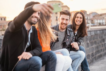 Photo for Group of young friends having fun sitting outdoors and taking memories with their smartphones at sunset - people and technology lifestyle concept - main focus on the brunette girl on the right - Royalty Free Image