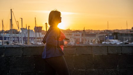Photo for Plus size woman exercising jogging at the port - Young beautiful chubby girl running along the coast holding a smartphone and listening to music with earphones at sunset - Backlit silhouette - People and body positive lifestyle concept - Royalty Free Image