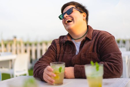 Selbstbewusste, nicht binäre Frau lächelt und scherzt mit Freunden am Tisch der Strandbar und trinkt Cocktails - Essen, Trinken, Bindung, Alkohol-Lifestyle-Konzept