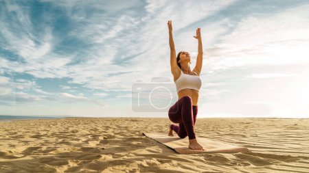 Full body three quarter view of serene female in sportswear - Girl stretching legs in Anjaneyasana while practicing yoga on the beach against a calm cloudy day at sunset