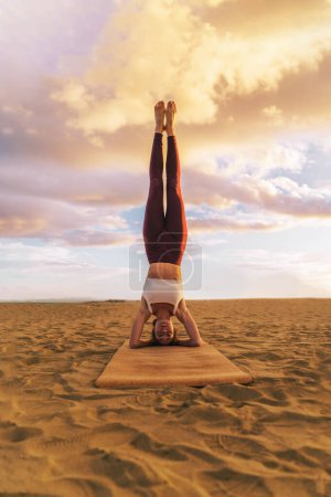 Téléchargez les photos : Belle jeune femme pratiquant le yoga sur la plage au coucher du soleil, faire de l'exercice de la tête, salamba sirsasana - concept de mode de vie sain des gens - plan vertical avec espace de copie - en image libre de droit