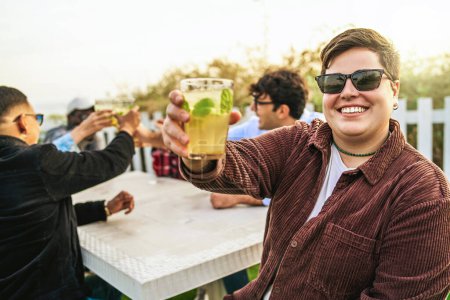 Photo for Multiethnic friends celebrating raising mojito cocktail glasses having fun together - happy people raising glasses together main focus on a non binary woman smiling, looking and toasting at the camera - Royalty Free Image