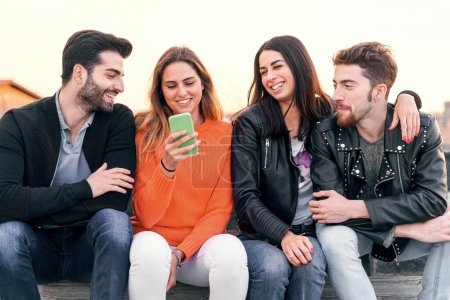 Happy group of trendy young people laughing and chatting sitting in a bench at sunset using a cellphone. Millennial friends having fun together outdoor - people, social network lifestyle concept