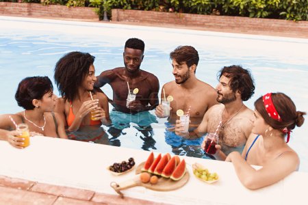 Téléchargez les photos : Multiethnic Group of Friends Relaxing in a Pool with Drinks and Fruit - A group of friends, including young adults of various ethnicities are enjoying a summer day drinking cold beverages - lifestyle - en image libre de droit