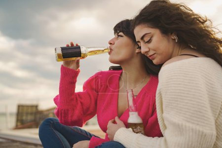 Two young caucasian women sit on a city wall, drinking Mexican beers and embracing each other, one with smooth hair, the other with curly hair, while a cloudy and gray sky serves as a background