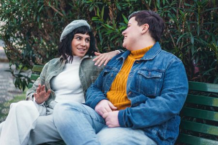 Photo for Two friends, a brunette and a curvy person of non-binary gender, are seen sitting on a park bench and having a conversation, both smiling - Perfect for illustrating themes of friendship, diversity, and lifestyle concepts. - Royalty Free Image