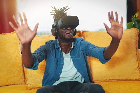 Téléchargez les photos : Young African American man sitting on couch, enjoying VR experience and exploring the metaverse. Perfect stock image for technology and gaming concepts - en image libre de droit