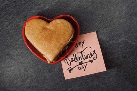 Photo for A heart-shaped pancake in a red heart-shaped saucer and a sticky note with Valentine's Day wishes left on the kitchen table - Royalty Free Image