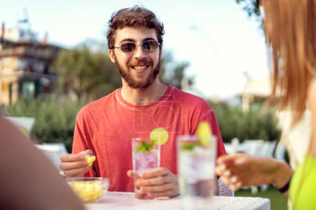 Foto de A young man is sitting at an outdoor pub table, smiling and having a great time with his friends (off-camera). He is dressed in a red t-shirt and holding a cocktail, teasing an appetizer - Imagen libre de derechos
