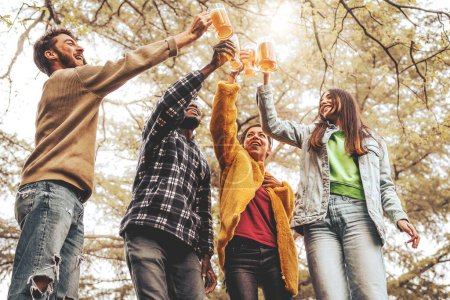 Photo for A group of four young adults standing and toasting with beer glasses raised high. Filled with joy and excitement as they celebrate in the beautiful countryside, greenery and bright sunshine - Royalty Free Image