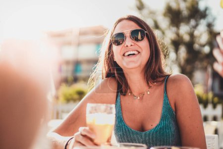 Foto de Chica joven sonriente en gafas de sol en la mesa al aire libre con amigos. Beber una bebida fresca en un día soleado de verano, con el sol creando un efecto de destello cálido. Amigos visibles pero no en el marco - Imagen libre de derechos