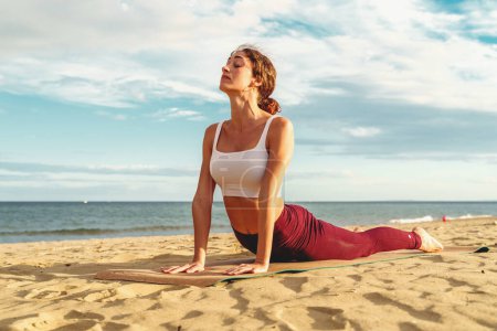 Foto de A young woman practices yoga on a mat positioned at the beach with the ocean horizon in the background. She is in the "Upward Facing Dog" pose, lifting her chest and straightening her back - Imagen libre de derechos
