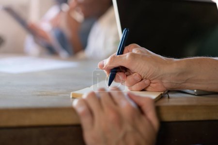 Photo for A close-up shot of a female hand holding a pen and writing in a notebook in an office setting. Other people are seated on the same desk, but are blurred in the background - Royalty Free Image