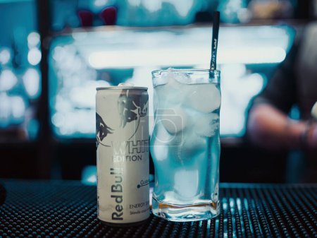 Téléchargez les photos : An editorial illustration showing a can of RedBull White Edition on a bar counter next to a glass with ice and a straw. The depth of field is very narrow with lights in the background of a pub or bar. - en image libre de droit