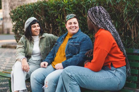 Foto de Tres mujeres, dos caucásicas (una morena, una curvilínea y no binaria) y una africana con extensiones de rastas en el pelo, se sientan juntas en un banco del parque, conversando - Imagen libre de derechos