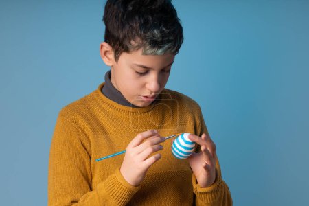 Foto de Sweet 10-year-old caucasian boy painting a white Easter egg with a paintbrush on a blue background. Great for Easter-themed designs or illustrating children's crafts and activities - Imagen libre de derechos