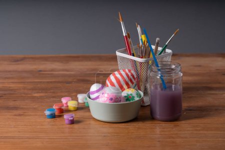 Photo for A collection of hand-decorated Easter eggs in a bowl, surrounded by paint jars, brushes, and a pencil holder on a wooden table. Ideal for Easter arts and crafts, DIY projects, and seasonal decoration - Royalty Free Image