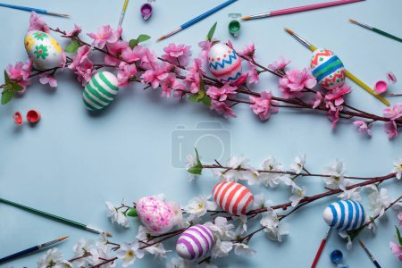 Foto de Fondo de temática pascual con huevos decorados, flores de cerezo y almendras dispuestas sobre fondo azul pastel, con espacio para el texto. También cuenta con pinceles y colores tempera - Imagen libre de derechos