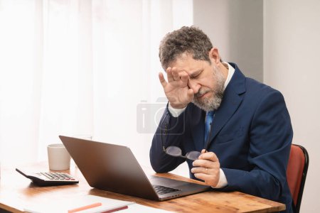 Téléchargez les photos : Un homme d'affaires fatigué d'âge moyen en costume bleu et cravate est assis à son bureau avec ses lunettes dans une main, se frottant les yeux avec l'autre. Utilisation prolongée d'un ordinateur et problèmes de vision liés à l'âge. - en image libre de droit