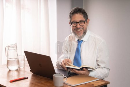 Foto de Confident mature businessman in formal businesswear sitting at his desk in the office. He looks at the camera with a smile, holding an agenda and a laptop. Perfect for corporate and business-themed designs - Imagen libre de derechos