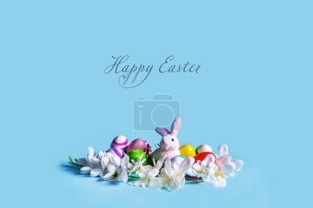 Foto de La tarjeta de Pascua presenta un diseño minimalista con un fondo azul pastel y una decoración hecha de flores, huevos decorados y un lindo conejito. El centro de se deja en blanco para el mensaje de la Feliz Pascua - Imagen libre de derechos