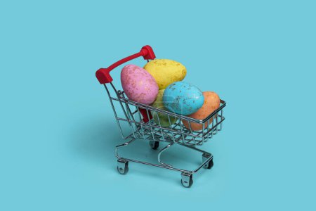 Foto de Un carrito de compras lleno de coloridos huevos de Pascua decorados con patrones divertidos y vibrantes sobre un fondo azul pastel. Perfecto para promociones de Semana Santa, tarjetas de felicitación y publicaciones en redes sociales. - Imagen libre de derechos