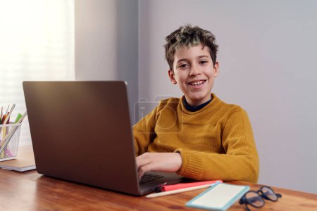 Foto de Un niño sonriente de 10 años de edad está sentado en un escritorio frente a una computadora portátil, con un suéter amarillo y gafas. El niño está estudiando, haciendo los deberes o aprendiendo en línea con la luz natural del día que entra por una ventana en el fondo. - Imagen libre de derechos