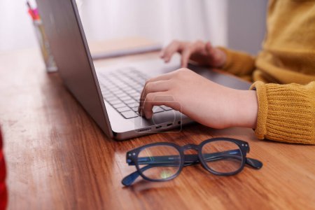 Téléchargez les photos : Une photo rapprochée des mains d'un enfant sur un ordinateur portable, avec le doigt déplaçant le curseur sur le trackpad. Le bureau en bois en arrière-plan a une paire de lunettes pliées éducation ou optométrie pédiatrique - en image libre de droit