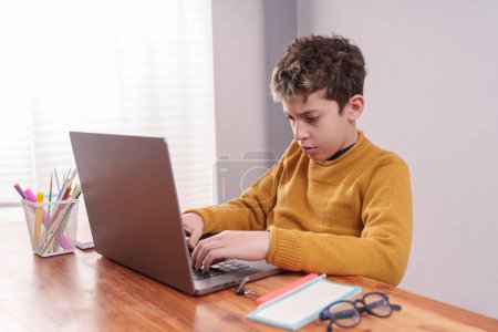 Téléchargez les photos : Vue latérale d'un jeune garçon assis à un bureau et tapant sur un ordinateur portable. Le garçon a une expression de concentration sur son visage pendant qu'il travaille. Au premier plan, il y a une paire de lunettes reposant sur le bureau - en image libre de droit