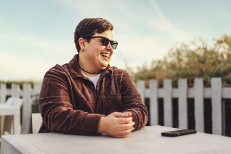 Téléchargez les photos : Une personne souriante de taille plus non binaire avec des lunettes de soleil dans un café extérieur, regardant ailleurs. Veste en velours marron, fond bleu ciel. - en image libre de droit