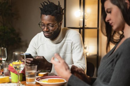 Téléchargez les photos : Un couple interracial - homme africain et femme caucasienne - fait l'expérience du phubbing dans un restaurant, alors qu'un partenaire est absorbé dans son smartphone, ignorant l'autre et causant une distance émotionnelle. - en image libre de droit