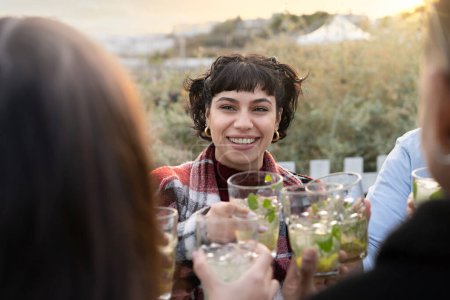Téléchargez les photos : Un groupe d'amis trinquant avec des cocktails frais dans un bar extérieur. Une femme brune souriante aux cheveux ondulés est à l'honneur. Fond flou avec végétation menant à la mer. - en image libre de droit