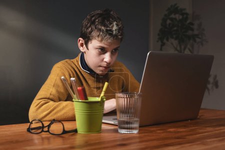 Foto de Niño enfocado de 10 años usando computadora portátil: en una habitación con poca luz, un niño se concentra en una pantalla de computadora portátil con luz de una ventana. Vasos, vaso de agua y porta bolígrafos en el escritorio de madera. - Imagen libre de derechos