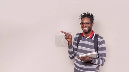 Foto de Retrato de un joven afroamericano con rastas cortas, mochila y cuadernos, sonriendo a la cámara y apuntando al espacio de copia. Estudio sobre fondo de luz cálida. - Imagen libre de derechos