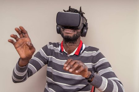 Téléchargez les photos : Homme afro-américain avec de courtes dreadlocks portant un casque de réalité virtuelle avec écouteurs intégrés, mains mobiles et geste dans la métaverse. - en image libre de droit