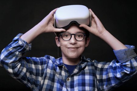Foto de Un niño caucásico de 12 años con gafas y una camisa a cuadros levanta sus auriculares VR, apoyándolos sobre su cabeza, mientras mira a la cámara y sonríe. El fondo es negro, destacando la emoción del niño por la tecnología de realidad virtual. - Imagen libre de derechos