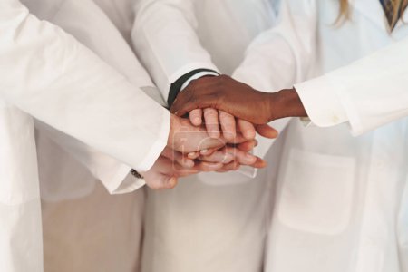 Foto de Un primer plano de un grupo de personas con batas de laboratorio, uniendo sus manos en un gesto de trabajo en equipo y colaboración, caras no visibles. - Imagen libre de derechos