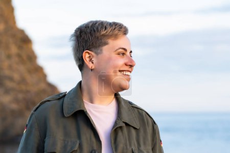 Photo for Portrait of a non-binary individual with short hair, slightly curvy, smiling by the sea. They are wearing a pink shirt and a green military jacket, looking away from the camera. - Royalty Free Image