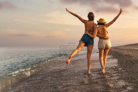 Téléchargez les photos : Deux jeunes amis, l'un portant un chapeau de paille, marchent sur la plage au coucher du soleil. Ils lèvent la main dans des gestes joyeux, exprimant amour et amitié. Concepts : amitié, vacances, désir d'errance. - en image libre de droit