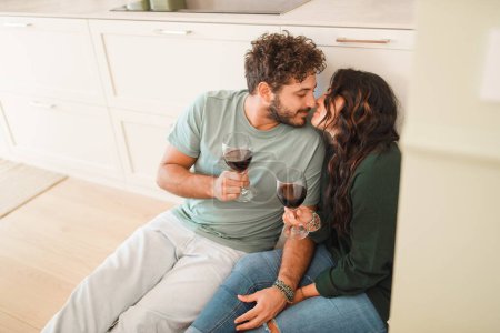 Photo for Intimate Moment: Young married couple sitting on the kitchen floor, holding wine glasses, hand in hand, sharing a kiss. Concept: love, togetherness, romance. - Royalty Free Image