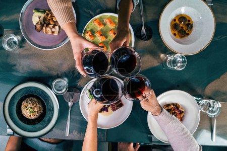 Foto de Vista aérea de cuatro amigos haciendo un brindis con vino tinto sobre una mesa de restaurante de fusión con varios platos - Imagen libre de derechos