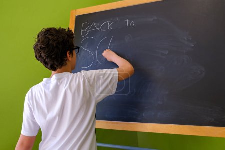 Foto de Vista posterior de un joven colegial escribiendo 'Back to School' con tiza blanca en una pizarra verde del aula - Imagen libre de derechos